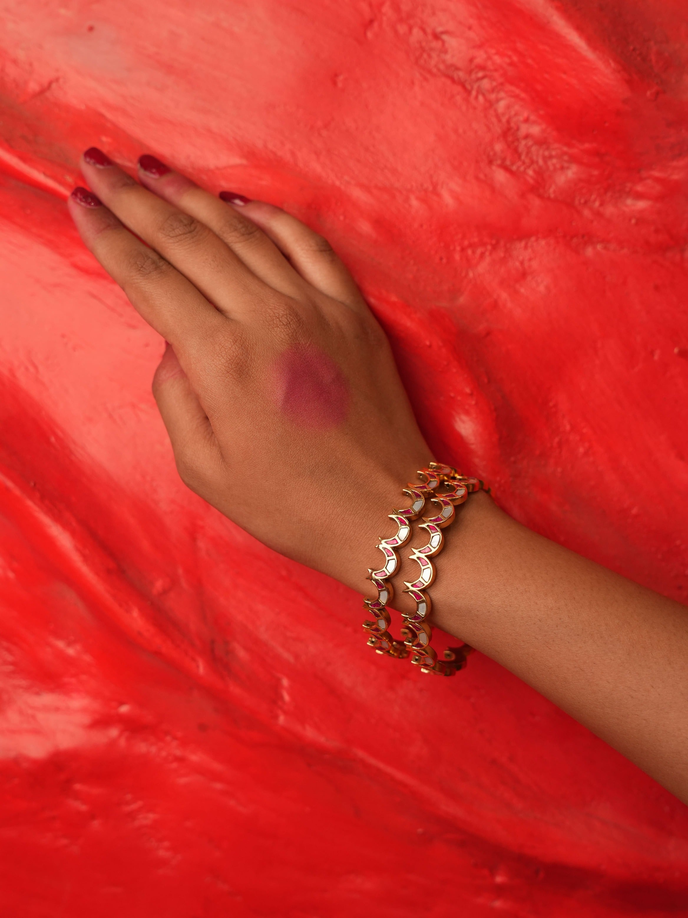 A close-up of a person's hand and wrist wearing a gold-toned multi-layered bangle bracelet, resting on a textured bright red surface.